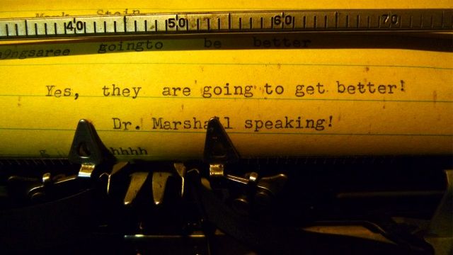 Inside the pharmacy, old equipment, medicine scales, and supplies are stacked on the counters and along the walls. In the back area where prescriptions were once filled, there are boxes of ancient Trojans and tins of pomade manufactured in Brooklyn. Here, a typewriter on the counter displays an optimistic message.